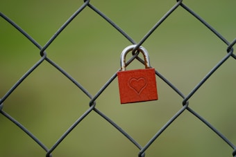 A red padlock with a heart engraving hangs on a wire fence, set against a blurred green background.
