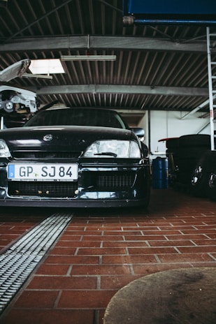 A black car is parked inside a garage with a red brick floor. The car has a visible license plate with the letters GP and SJ 84. In the background, the garage has a metal roof, and a partially visible hoisted car can be seen. There are various tools and equipment, including tires stacked to the side.