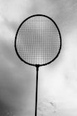 A silhouetted tennis racket is held against a cloudy sky, with the strings clearly visible.
