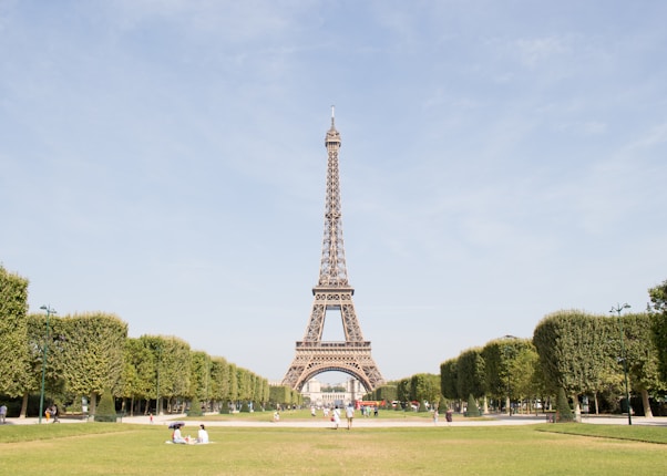 The Eiffel Tower stands prominently in the center of a well-manicured park with lush green lawns and evenly spaced trees. A few people are visible in the park, enjoying a sunny day.