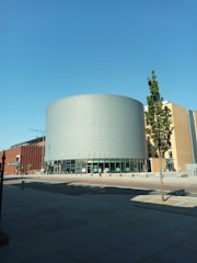 A modern building with cylindrical architecture featuring a sign for the University of Manchester. The facade includes glass windows and metal cladding. There is a small tree in the foreground and a clear blue sky above.