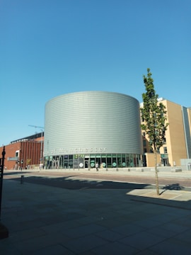 A modern building with cylindrical architecture featuring a sign for the University of Manchester. The facade includes glass windows and metal cladding. There is a small tree in the foreground and a clear blue sky above.