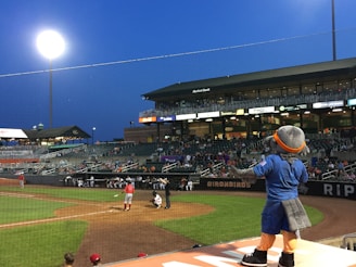 A baseball stadium under a clear evening sky with bright floodlights illuminating the field. Fans are seated in the stands, observing the ongoing game. A baseball player wearing a red uniform is preparing to bat, while the catcher and umpire are in position. A costumed mascot is seen in the foreground, dressed in blue and orange, engaging with the crowd.