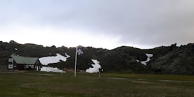 A solitary house with a green roof is positioned in a vast grassy landscape. A flagpole stands prominently, flying the national flag of Iceland. Snow patches cover parts of the surrounding dark rocky hills under a cloudy sky.