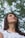 A young woman gazes upward, surrounded by blurred green foliage in the background. Her expression is serene, with a slight smile and relaxed demeanor. She is wearing a white top with intricate patterns and long dark hair falls over her shoulders.