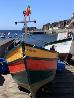 A colorful fishing boat with yellow, red, and green stripes is positioned on a dock. The boat has a small flag on top with a cross-like structure behind it. In the background, several other boats are floating on calm, blue water. Rugged cliffs and a concrete harbor structure are visible under a clear blue sky.