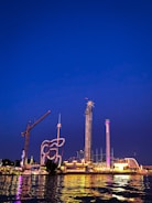 An amusement park is illuminated against the night sky, featuring various thrill rides such as a tall drop tower, a looping roller coaster, and a brightly lit carousel. The reflection of the lights dances on the water in the foreground.