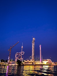 An amusement park is illuminated against the night sky, featuring various thrill rides such as a tall drop tower, a looping roller coaster, and a brightly lit carousel. The reflection of the lights dances on the water in the foreground.