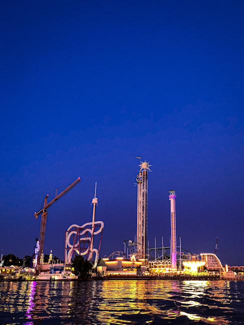 An amusement park is illuminated against the night sky, featuring various thrill rides such as a tall drop tower, a looping roller coaster, and a brightly lit carousel. The reflection of the lights dances on the water in the foreground.
