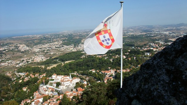 A picturesque landscape features a panoramic view over a hilly area with a mix of urban and natural surroundings. A prominent white flag with a coat of arms is flying on a pole in the foreground, capturing attention with its vivid design. Below, a historic village with white buildings topped with terracotta roofs is nestled among lush green trees, offering a blend of nature and architecture. The expansive view extends to the distant horizon, revealing a clear blue sky meeting the edge of the land.