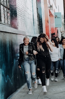A group of people walking together along a sidewalk beside a wall with colorful graffiti. They are smiling and seem to be enjoying each other's company. The group appears diverse and dressed in casual attire, with some wearing jeans and jackets.
