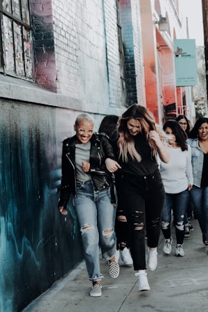 A group of people walking together along a sidewalk beside a wall with colorful graffiti. They are smiling and seem to be enjoying each other's company. The group appears diverse and dressed in casual attire, with some wearing jeans and jackets.