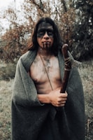 A person stands in a natural outdoor setting surrounded by trees, holding a wooden staff with a stone blade. The individual is wrapped in a dark green cloak and has black face paint, including stripes across the face.