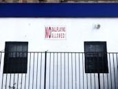 A white wall features a prominent sign reading 'No Ball Playing Allowed.' The sign is red and white, positioned between two barred windows. A black metal fence extends across the lower portion of the image. A security camera is mounted above to the right.
