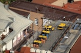 A rooftop terrace featuring several tables and chairs, primarily in yellow and blue colors, is surrounded by metal railings. String lights are hung across the area, and the rooftop is built atop a multi-story brick building with a red sloped roof. The adjacent structure has light-colored walls and a metallic roof. There are also decorative plants and a visible corridor with laundry hanging.