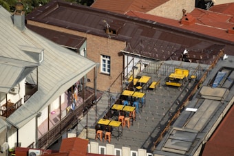 A rooftop terrace featuring several tables and chairs, primarily in yellow and blue colors, is surrounded by metal railings. String lights are hung across the area, and the rooftop is built atop a multi-story brick building with a red sloped roof. The adjacent structure has light-colored walls and a metallic roof. There are also decorative plants and a visible corridor with laundry hanging.