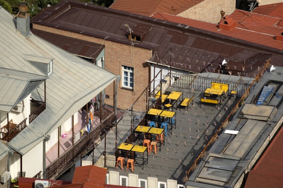 A rooftop terrace featuring several tables and chairs, primarily in yellow and blue colors, is surrounded by metal railings. String lights are hung across the area, and the rooftop is built atop a multi-story brick building with a red sloped roof. The adjacent structure has light-colored walls and a metallic roof. There are also decorative plants and a visible corridor with laundry hanging.