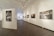 An art gallery interior with various photographs and paintings hanging on the white walls. The flooring is wooden, and overhead track lighting illuminates the artworks. The space is spacious and well-lit, creating an inviting atmosphere for viewing art.