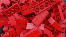 A large pile of red plastic interlocking building bricks, some lying flat while others are stacked at angles. Various sizes and shapes are visible, with the classic cylindrical connectors prominent on their surfaces.