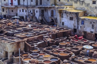 A traditional tannery with numerous circular stone vats filled with various shades of liquid. Workers are seen handling hides across the area, surrounded by old, rustic buildings with worn exteriors. Some of the vats contain hides being soaked in colorful liquids, while others are empty or filled with water.