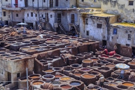 A traditional tannery with numerous circular stone vats filled with various shades of liquid. Workers are seen handling hides across the area, surrounded by old, rustic buildings with worn exteriors. Some of the vats contain hides being soaked in colorful liquids, while others are empty or filled with water.