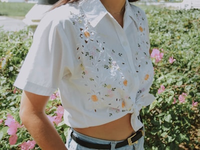 A person wearing a white shirt with floral embroidery, tied at the waist. The shirt features bright yellow and green details. The background consists of vibrant pink flowers and lush green foliage on a sunny day.