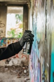 A gloved hand is holding a spray paint can close to a wall covered in colorful graffiti. The setting appears to be an abandoned building, with unfinished and rough textures visible in the wall and surroundings. Natural light comes through a large window in the background, partially illuminating the scene.