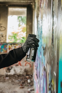 A gloved hand is holding a spray paint can close to a wall covered in colorful graffiti. The setting appears to be an abandoned building, with unfinished and rough textures visible in the wall and surroundings. Natural light comes through a large window in the background, partially illuminating the scene.