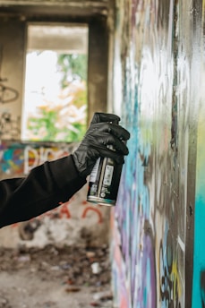 A gloved hand is holding a spray paint can close to a wall covered in colorful graffiti. The setting appears to be an abandoned building, with unfinished and rough textures visible in the wall and surroundings. Natural light comes through a large window in the background, partially illuminating the scene.