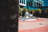 A modern urban plaza featuring a central fountain with a contemporary metal sculpture. Surrounding the fountain are brick and concrete walkways, and the background includes a building with reflective glass windows. Lush greenery and hanging flower baskets add color and vibrancy to the scene.
