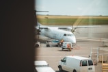 A white airplane is stationed on a tarmac at an airport. Various ground vehicles and equipment, including a white van and a baggage cart, are present near the aircraft. The aircraft appears to be undergoing service or preparation. There are open fields in the background under a clear sky.