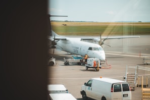 A white airplane is stationed on a tarmac at an airport. Various ground vehicles and equipment, including a white van and a baggage cart, are present near the aircraft. The aircraft appears to be undergoing service or preparation. There are open fields in the background under a clear sky.