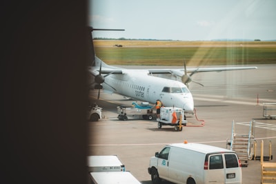 A white airplane is stationed on a tarmac at an airport. Various ground vehicles and equipment, including a white van and a baggage cart, are present near the aircraft. The aircraft appears to be undergoing service or preparation. There are open fields in the background under a clear sky.