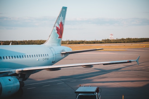 An airplane is parked on an airport tarmac with a wing prominently visible. The aircraft's tail features a distinctive logo of a red maple leaf on a blue and white background. The surrounding area is open with distant trees and a cloudy sky.
