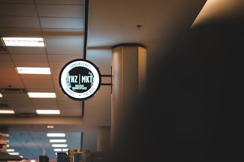 A dimly lit interior features a circular sign for a marketplace, highlighting sandwiches, soups, and salads. The sign reads 'YHZ MKT' and 'Halifax Marketplace' and is mounted on a pole. The ceiling has several fluorescent lights, and the overall ambiance is subdued.