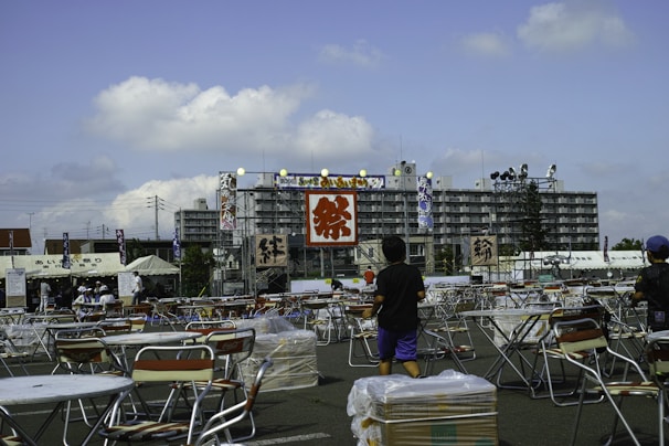 An outdoor festival setting with numerous foldable tables and chairs arranged in rows on a paved area. A large building serves as a backdrop, adorned with banners featuring Japanese characters. Two children are walking among the furniture, and several tents are set up, suggesting preparation for a public event. The sky is partly cloudy, adding a bright and open atmosphere.