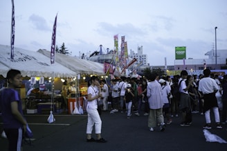 A bustling outdoor market or festival scene with numerous people walking around and interacting. Several tents with food and goods are set up, adorned with colorful signs and flags. Bright lights illuminate the various stalls as the evening seems to be approaching. In the background, buildings and a sign for 'COOP Sapporo' are visible.