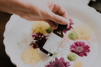 A hand holds a small brown glass bottle above a white dish filled with colorful flowers, including pink and yellow blooms. The flowers are arranged in a decorative manner, creating a visually pleasing aesthetic.