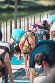 Several people are participating in an outdoor yoga class, performing what appears to be a forward bend pose. The setting is bright with a natural background, likely near a park or body of water. Participants are using yoga mats, and they are dressed in athletic wear.