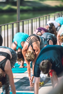 Several people are participating in an outdoor yoga class, performing what appears to be a forward bend pose. The setting is bright with a natural background, likely near a park or body of water. Participants are using yoga mats, and they are dressed in athletic wear.