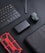 A flat lay arrangement features various tech gadgets on a dark surface. A black wireless keyboard is placed diagonally, accompanied by a black portable charger with a subtle brand logo. Next to it, there is a compact action camera with a small display screen showing a preview. A red and black rugged smartphone case catches attention with its bold design. A black case or bag is partially visible in the frame.