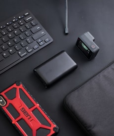 A flat lay arrangement features various tech gadgets on a dark surface. A black wireless keyboard is placed diagonally, accompanied by a black portable charger with a subtle brand logo. Next to it, there is a compact action camera with a small display screen showing a preview. A red and black rugged smartphone case catches attention with its bold design. A black case or bag is partially visible in the frame.