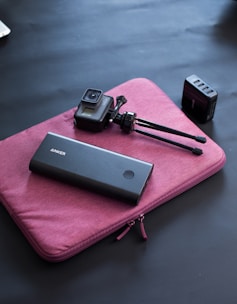 A black power bank, a small camera with a tripod, and a black multi-port charger are placed on a purple laptop sleeve on a dark surface.