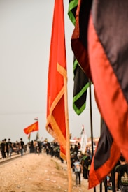 A group of people walking on a dirt path alongside a railway track carrying various flags. The flags are prominently red and black, with some having green accents. The sky is overcast, giving the image a muted light.