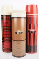 Three vintage thermos bottles in varied designs are arranged side by side. The left thermos features a red and black plaid pattern, while the center one is solid brown with a stripe design. The right thermos showcases a red and black vertical striped pattern. All three have cream-colored tops.
