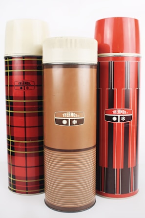 Three vintage thermos bottles in varied designs are arranged side by side. The left thermos features a red and black plaid pattern, while the center one is solid brown with a stripe design. The right thermos showcases a red and black vertical striped pattern. All three have cream-colored tops.