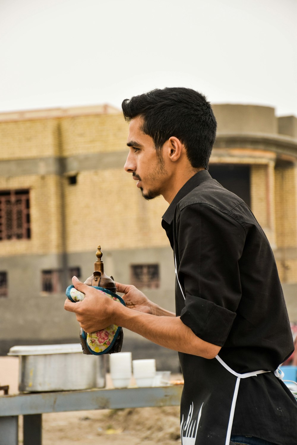 Mann mit schwarzem Hemd und Schürze steht und hält eine Flasche in der Hand