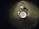 A metal sink drain surrounded by water droplets on a dark surface. The area around the drain has a reflective appearance with scattered droplets creating a glistening effect.