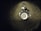 A metal sink drain surrounded by water droplets on a dark surface. The area around the drain has a reflective appearance with scattered droplets creating a glistening effect.