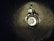 A metal sink drain surrounded by water droplets on a dark surface. The area around the drain has a reflective appearance with scattered droplets creating a glistening effect.
